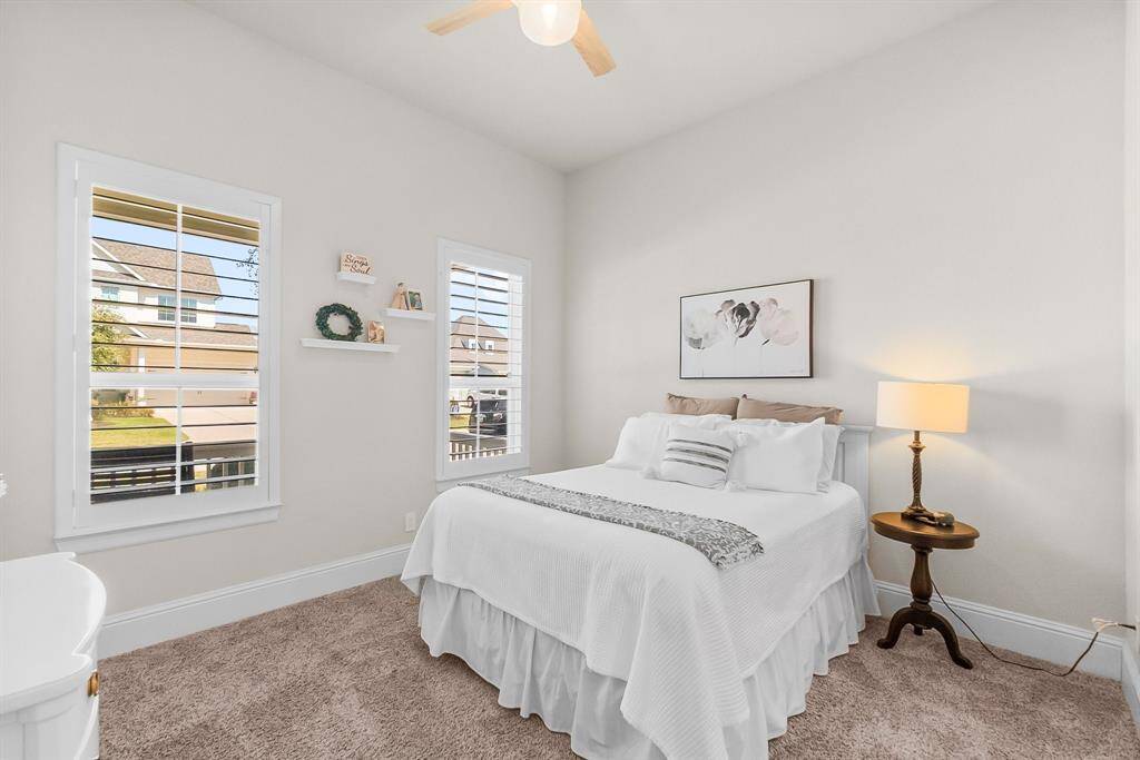 Second bedroom features soft carpeting, light walls, and ample natural light from two windows with shutters.