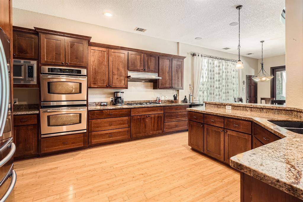 This kitchen features rich wooden cabinetry, granite countertops, and stainless steel appliances, including a double oven and microwave. The open layout includes a large island with a breakfast bar, pendant lighting, and a view into the dining area, creating a warm and inviting space for cooking and entertaining.