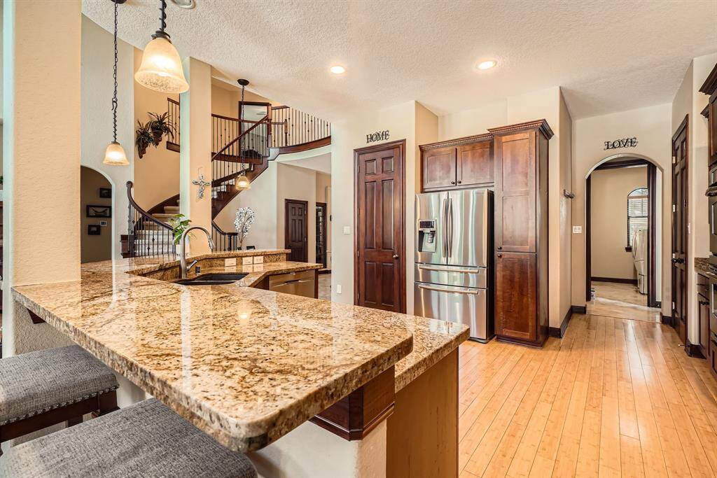 This photo showcases a spacious, modern kitchen with granite countertops, stainless steel appliances, and warm wood cabinetry. The open layout includes a stylish breakfast bar with pendant lighting and a view of a curved staircase, adding elegance to the space.
