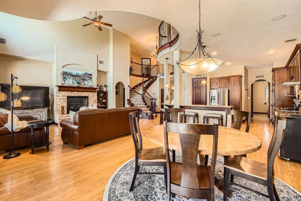 This spacious open-concept living area features a cozy stone fireplace, elegant spiral staircase, and modern kitchen with stainless steel appliances. The room is highlighted by high ceilings, a mix of warm wood tones, and a stylish dining area, creating a welcoming and luxurious atmosphere.