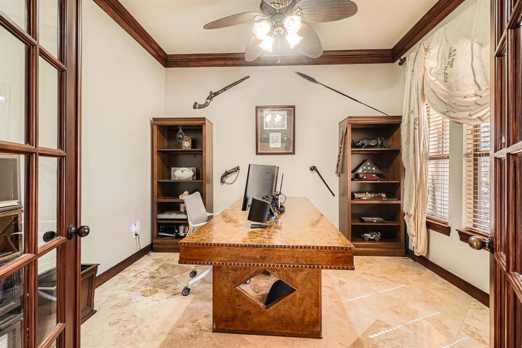 This is a home office with elegant French doors, featuring a large wooden desk, with shelving spaces and ample natural light from the windows. The room is accented with decorative items and has a ceiling fan for comfort.