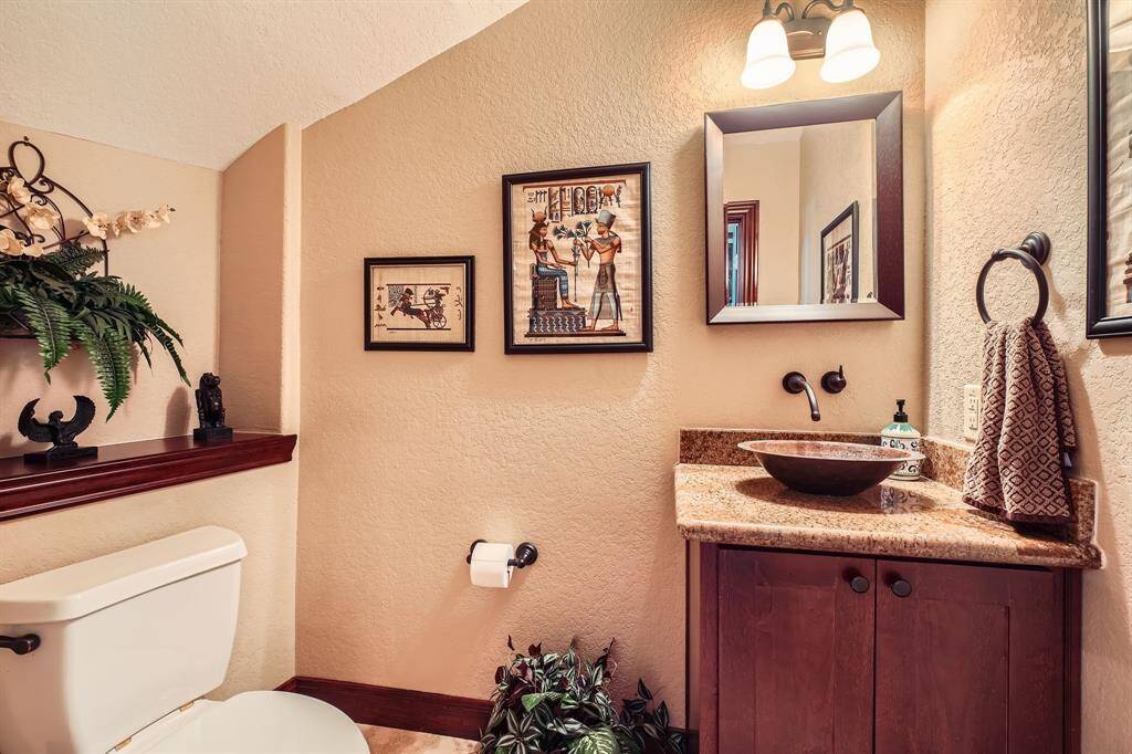 This bathroom features a warm, inviting design with textured walls and decorative artwork. It includes a stylish vessel sink on a granite countertop, dark wood cabinetry, and chic lighting. The space is accented with greenery and elegant decor elements, creating a cozy and sophisticated atmosphere.