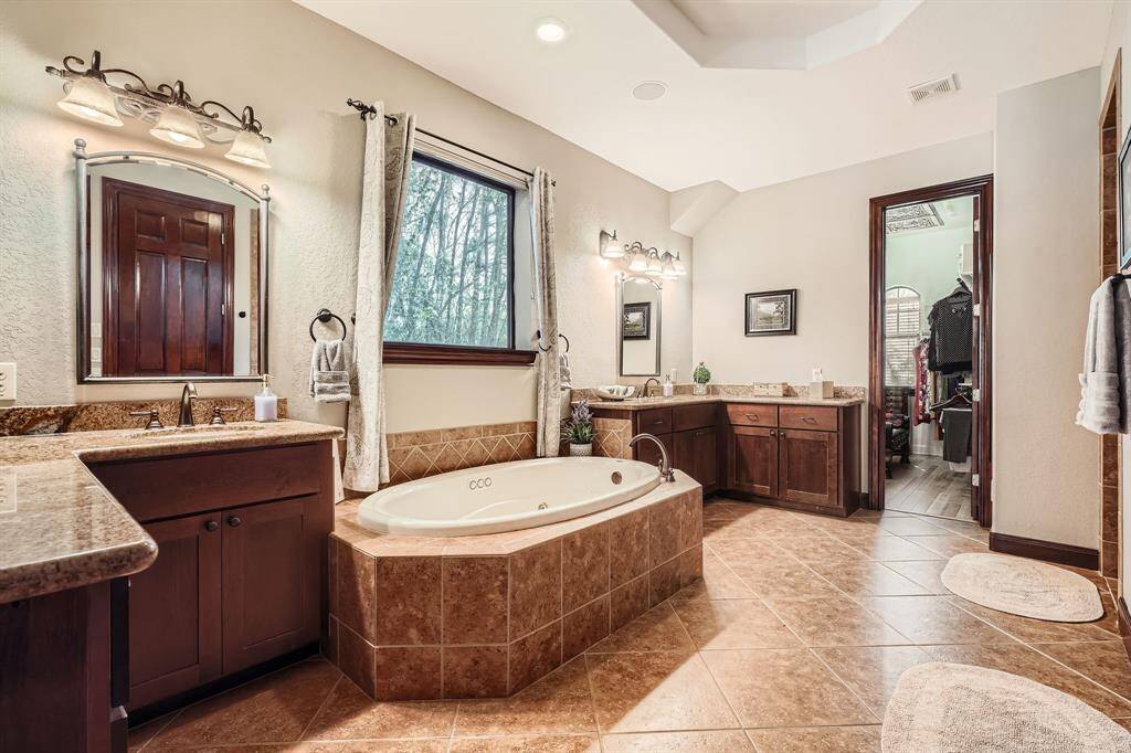 This spacious bathroom features a luxurious bathtub, dual sinks with granite countertops, elegant wooden cabinetry, and a large window offering natural light. The room is complemented by stylish tile flooring and leads into a walk-in closet.