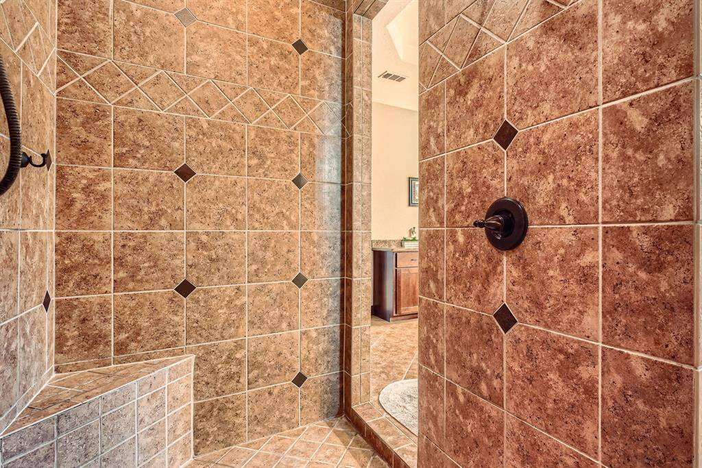This photo showcases a spacious, tiled shower with earthy tones and decorative accents, featuring a built-in bench and modern fixtures, leading into a well-lit bathroom area.