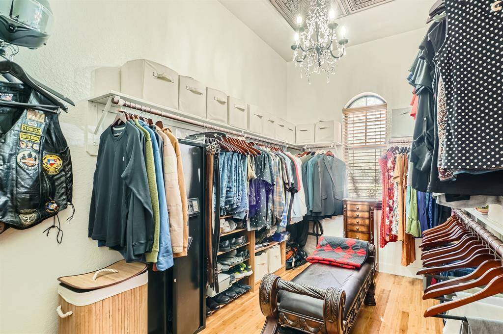 This spacious walk-in closet features ample hanging and shelf storage, hardwood floors, and a stylish chandelier, all illuminated by natural light from a window with wooden blinds.