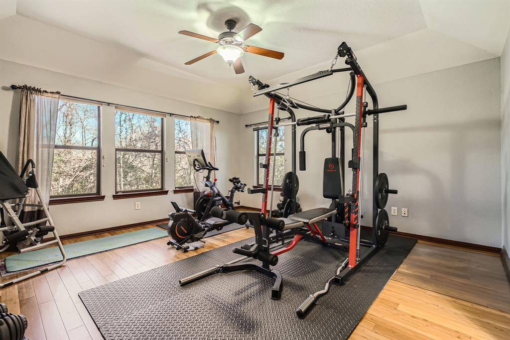 This room features a spacious home gym with various exercise equipment. Large windows provide plenty of natural light and a view of the surrounding greenery. The space has wood flooring and a ceiling fan for comfort.