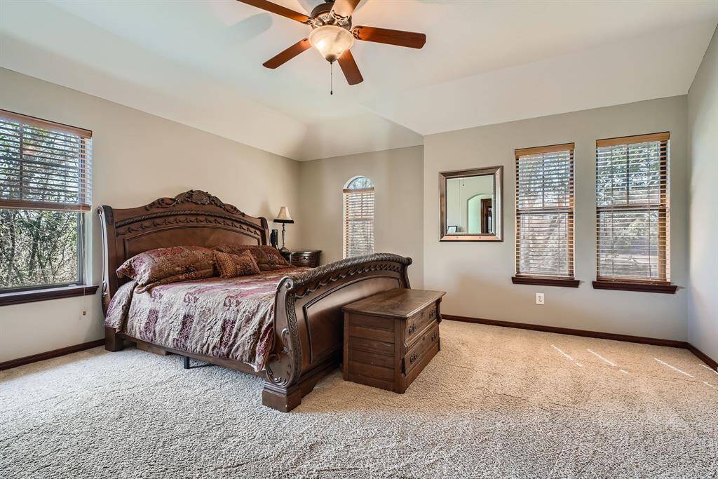 This spacious bedroom features a large, ornate wooden bed, neutral walls, and plush carpeting. It has ample natural light from three windows with wooden blinds and a ceiling fan for comfort.