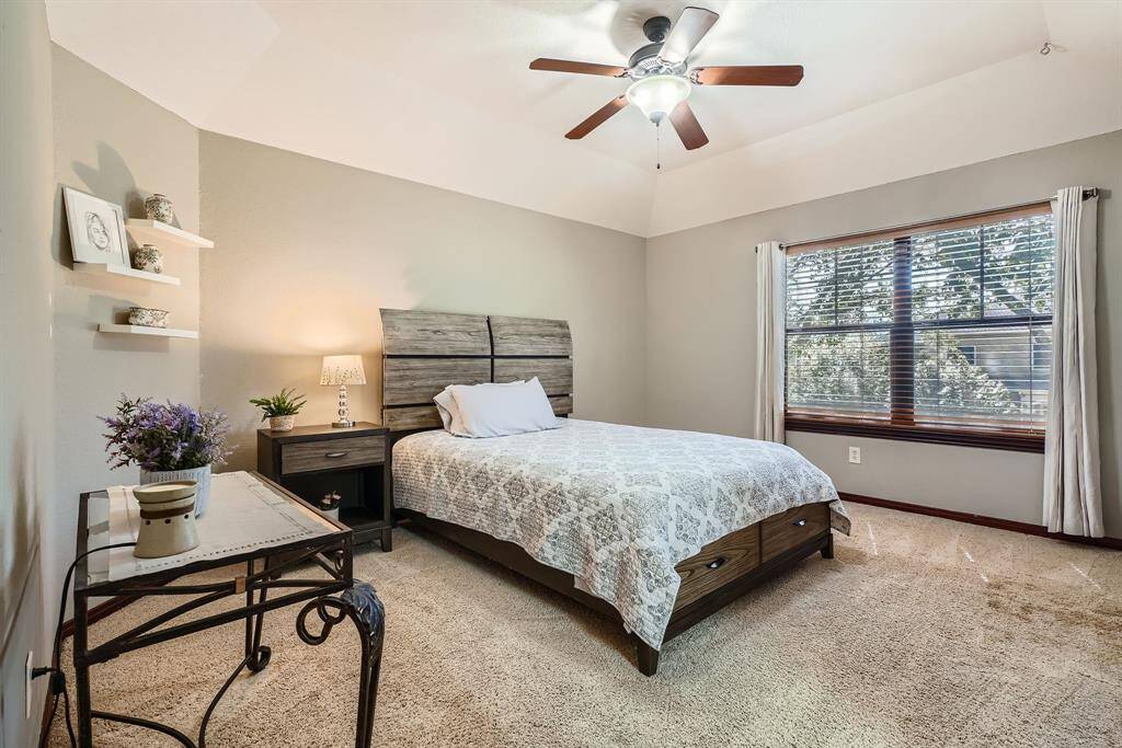This cozy bedroom features a comfortable and spacious area, a ceiling fan, and a large window with blinds and curtains. The neutral-toned walls and carpet create a warm atmosphere.