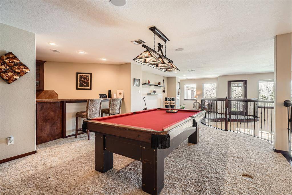 This inviting recreational room features a stylish pool table and a full kitchenette to the left, ambient lighting, and a cozy bar area with seating. The space is accented by neutral walls, carpet flooring, and offers ample natural light from large windows, creating an ideal spot for entertainment and relaxation.