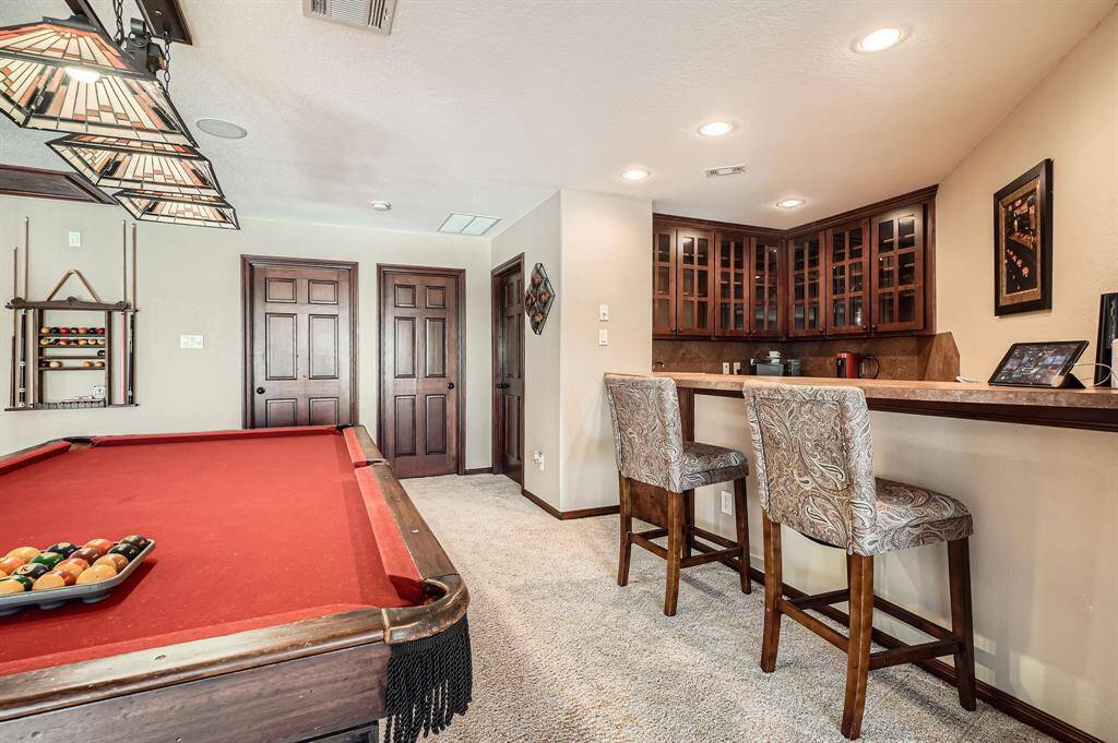 This photo showcases a cozy recreation room with a stylish bar area and a full kitchenette  featuring two high chairs, and ample cabinet storage with glass doors. The room has warm lighting and plush carpeting, creating an inviting atmosphere for entertainment.
