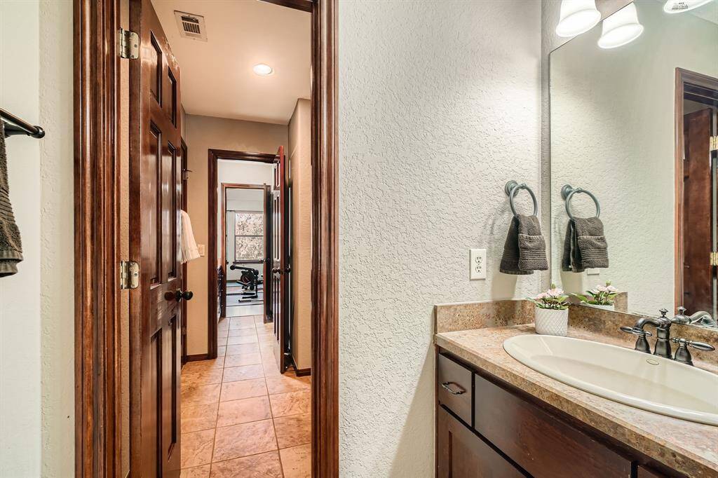 This photo shows a well-lit bathroom featuring a modern vanity with a stone countertop and under-mount sink. Adjacent is a hallway with tile flooring, leading to another room. Warm wood doors and trim add a cozy touch.
