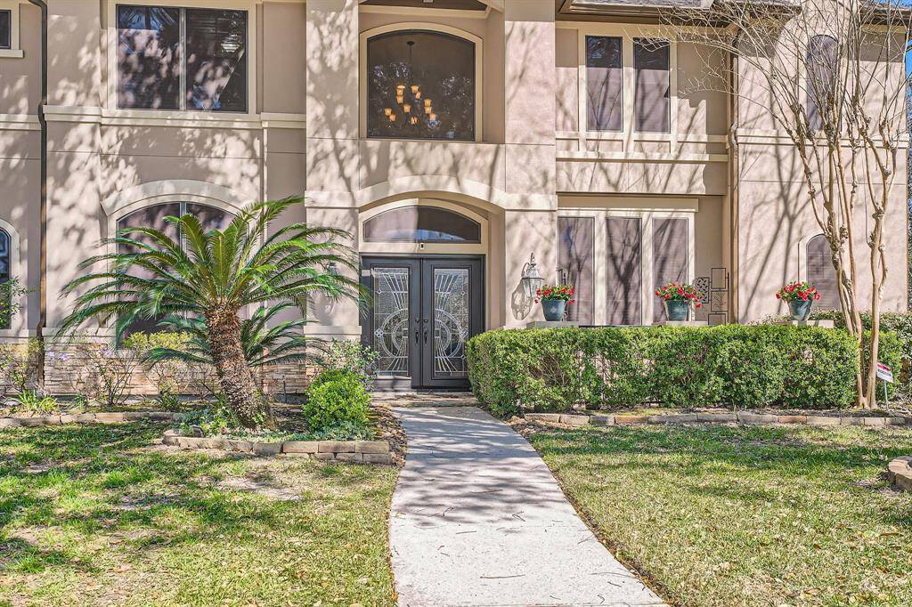 This elegant home features a grand entrance with decorative double doors, surrounded by well-maintained landscaping, including a small palm tree and vibrant potted flowers. The facade is accented by large windows and tasteful architectural details, creating an inviting and sophisticated curb appeal.