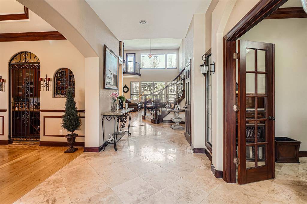 A spacious, elegant entryway with high ceilings, featuring a mix of wood and tile flooring. It includes a wrought iron wine cellar door, French doors leading to a study or office, and a view of the living area with large windows that allow natural light to fill the space. The staircase and tasteful decor add a touch of sophistication.