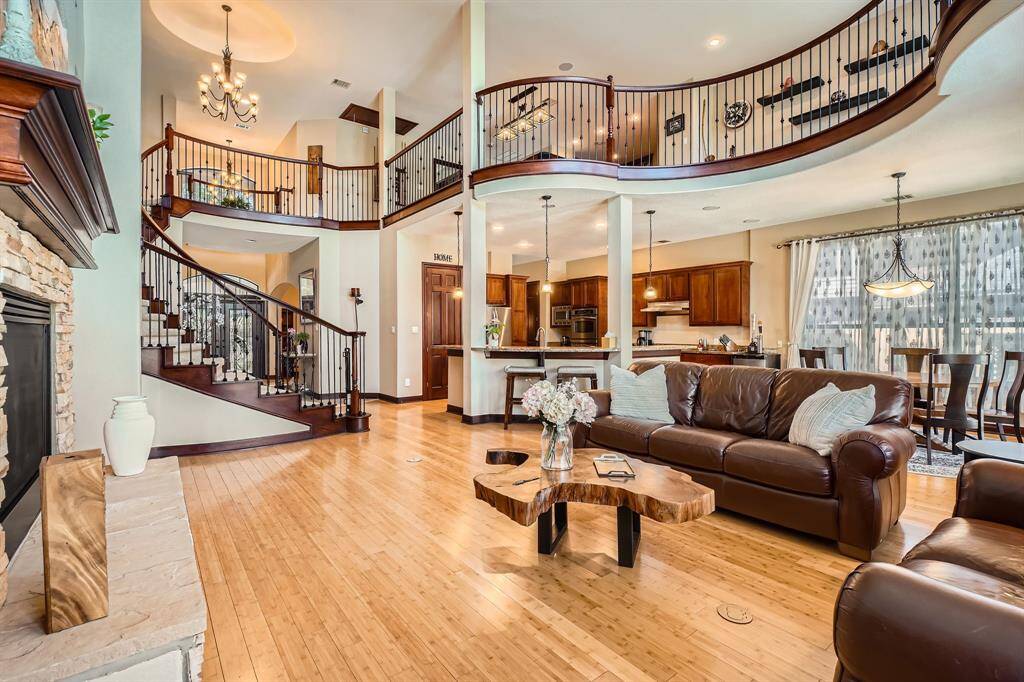 This spacious living room features an open floor plan with high ceilings and elegant wooden flooring. It includes a cozy seating area with leather sofas, a stone fireplace, and a unique wooden coffee table. The room seamlessly connects to a modern kitchen and dining area, with a stylish staircase leading to an upper level. Natural light floods in through large windows, enhancing the warm and inviting atmosphere.