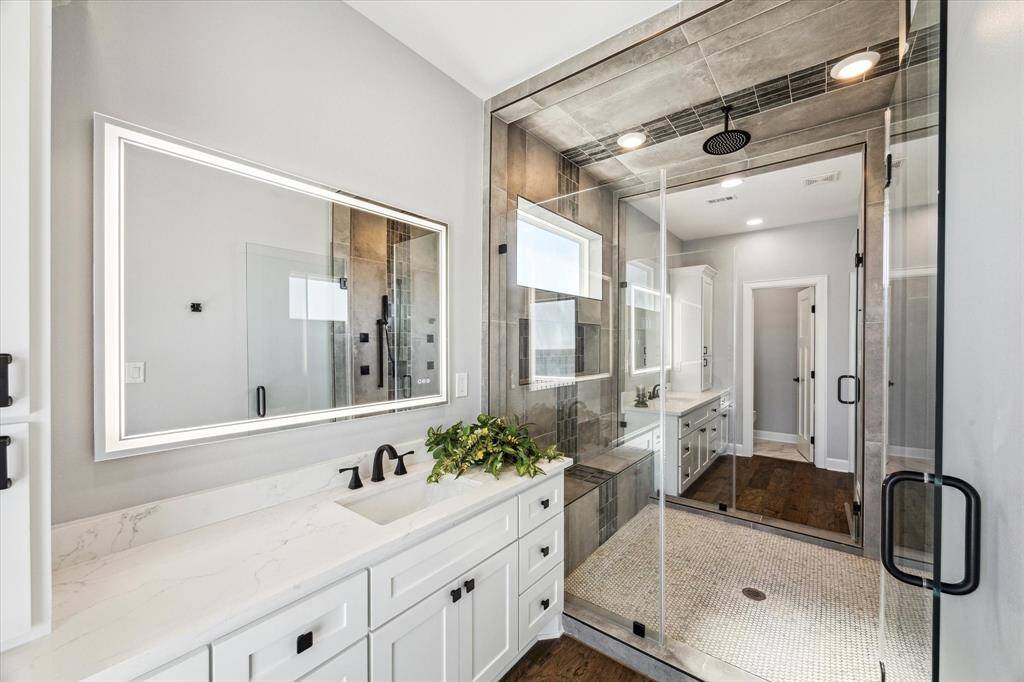 PRIMARY BATH WITH HIS AND HER VANITIES WITH BACK-LIT MIRRORS CONNECTED BY A WALKTHROUGH SHOWER WITH FLOOR TO CEILING TILE SURROUND AND A RAIN SHOWER HEAD.