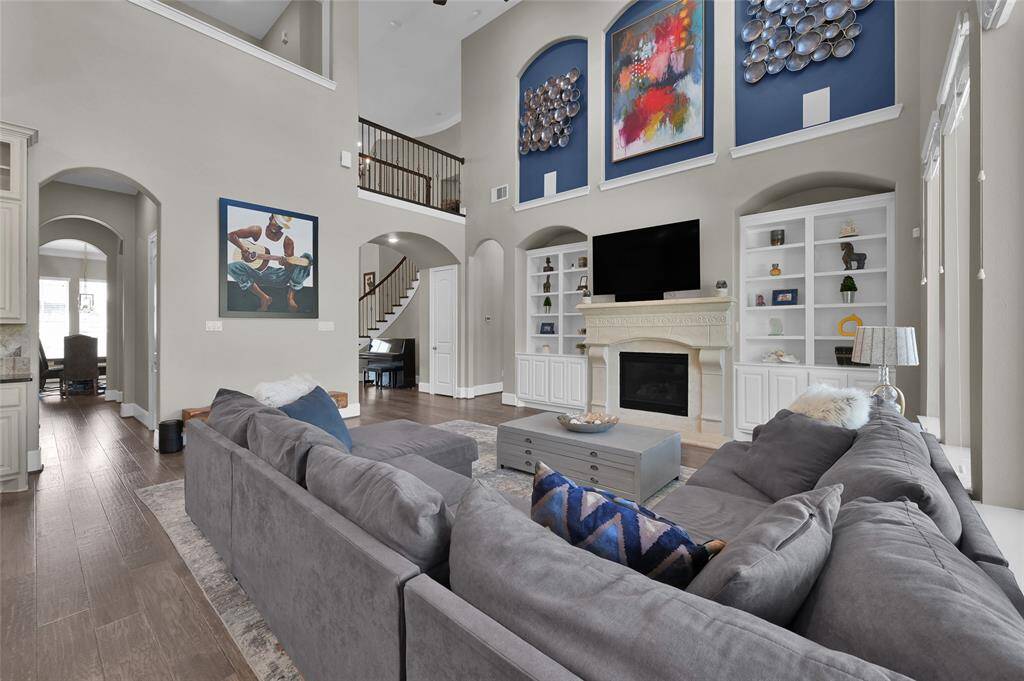 This spacious living room features high ceilings, abundant natural light, and elegant decor. It includes a cozy fireplace and built-in shelving,  perfect for relaxation and entertaining. The open layout leads to a dining area, enhancing the home's inviting atmosphere.