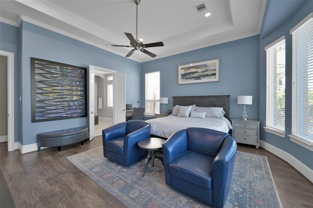 This spacious primary bedroom features a calming blue color scheme with ample natural light from multiple windows. The room also has wood floors, a ceiling fan, and access to an en-suite bathroom.