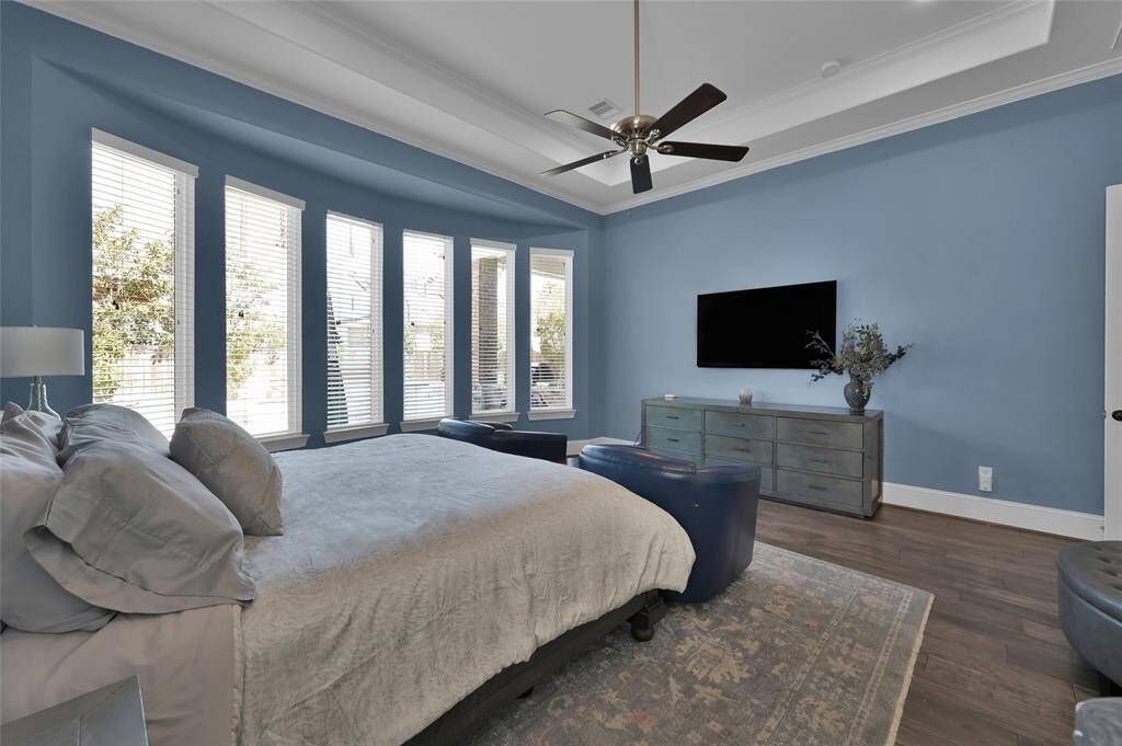 This spacious primary bedroom features a soothing blue color scheme, large windows for natural light, a ceiling fan, and a mounted TV.