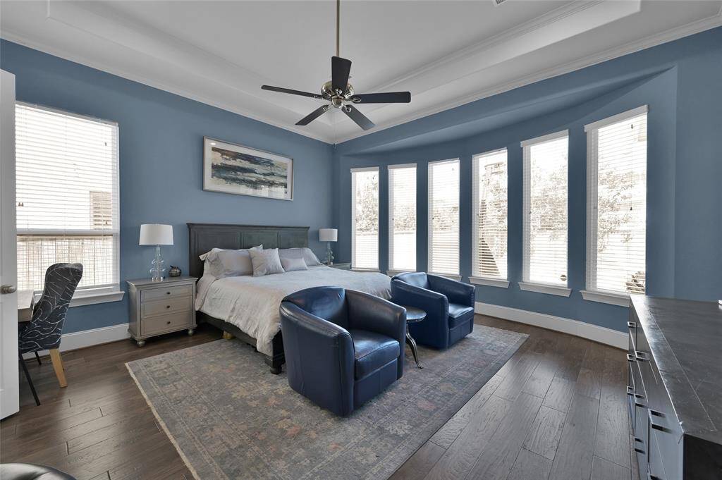 This spacious master bedroom features elegant blue walls, abundant natural light from large bay windows, and a stylish ceiling fan.
