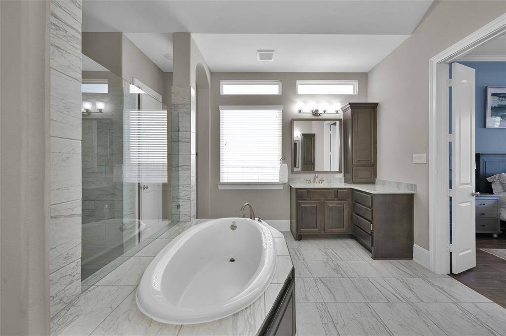 This photo features a modern bathroom with a luxurious soaking tub, a spacious walk-in shower, and elegant tile flooring. The room is bright, with natural light from high windows, and includes a well-appointed vanity with ample storage.