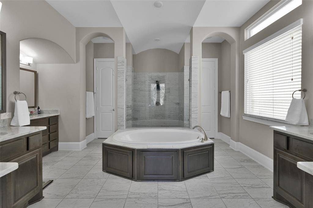 This bright and airy bathroom features a central soaking tub, elegant marble tile flooring, and dual vanities with ample storage. Large windows provide natural light, and the shower area is enclosed in glass, creating a luxurious and serene atmosphere.