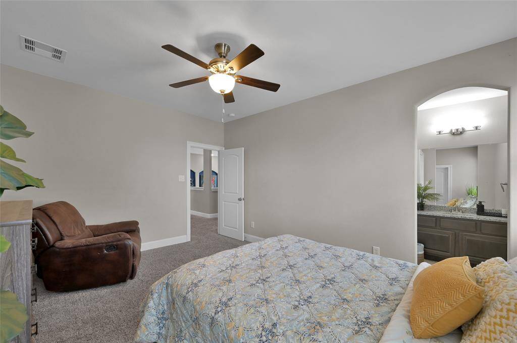 This bedroom features neutral walls, a ceiling fan, and plush carpeting. It includes a cozy seating area and a view into an en-suite bathroom with modern fixtures.