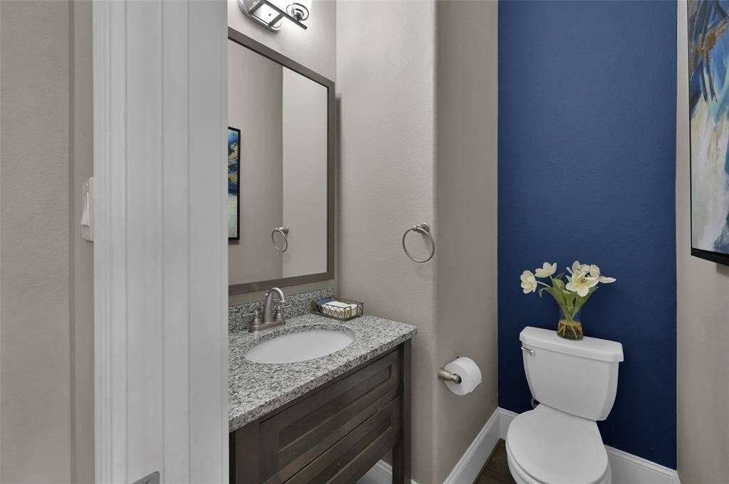 This is the half bath located downstairs and it is a compact, modern half-bathroom featuring a granite countertop with a single sink, a sleek mirror, and a toilet. The space is accented with a stylish blue wall and decorative artwork, creating a fresh and inviting atmosphere.