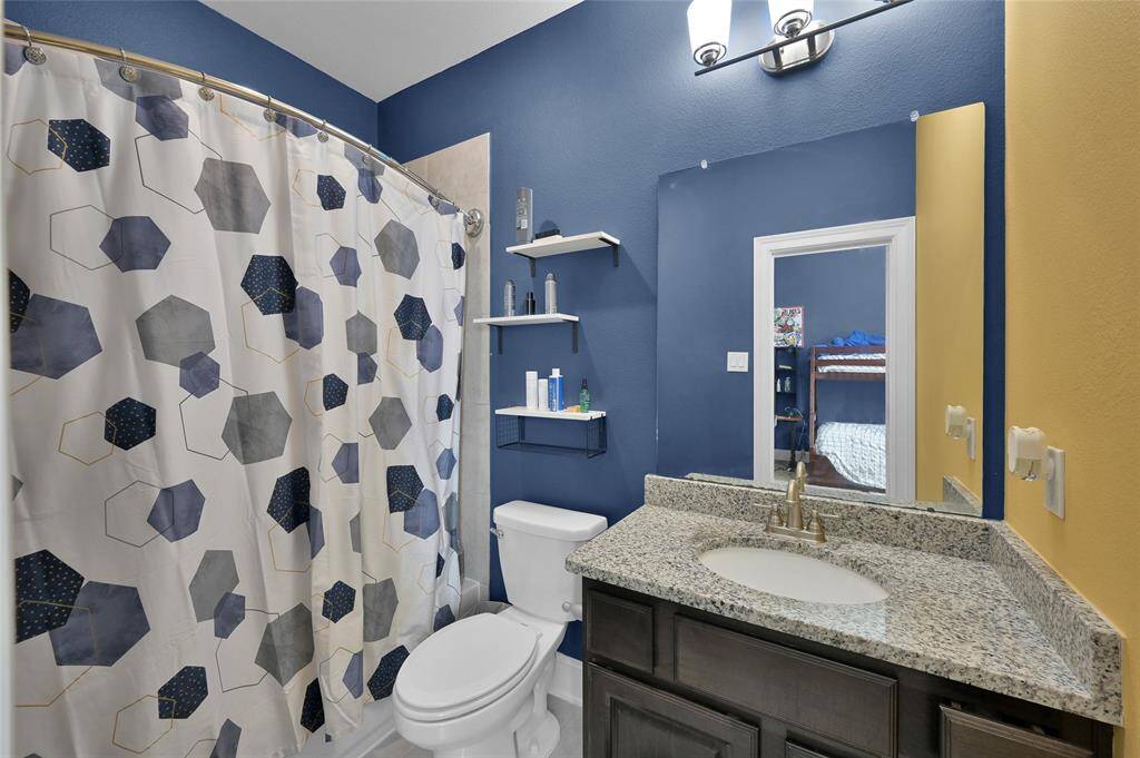 Another one of the bathrooms upstairs.  This features a modern design with a granite countertop, dark wood vanity, and a large mirror. The walls are painted in a bold blue, complemented by a geometric-patterned shower curtain. Shelving provides extra storage above the toilet, and contemporary lighting adds a stylish touch.