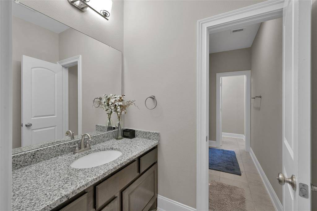 This is one of the modern bathrooms upstairs featuring a granite countertop with an undermount sink, sleek cabinetry, and a large mirror. The neutral walls complement the stylish fixtures, and the hallway beyond reveals a tiled floor with a blue rug, adding a touch of color.