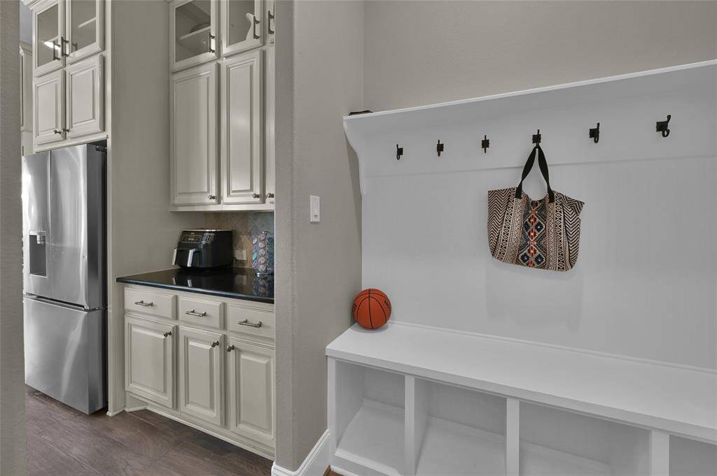This photo features a modern entryway with built-in white storage benches and coat hooks, adjacent to a kitchen area with stainless steel appliances and cream-colored cabinetry.