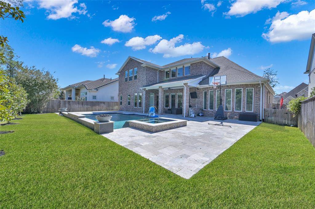 This spacious backyard features a modern two-story brick home with large windows, a well-maintained lawn, a paved patio area, and an inviting pool. Perfect for entertaining or relaxing outdoors.