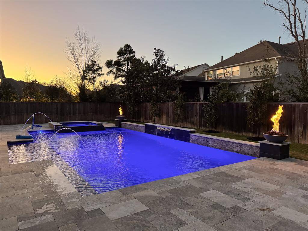 This photo showcases a spacious backyard featuring a modern swimming pool with vibrant lighting, adjacent spa, and elegant fire bowls. The pool is surrounded by a stone patio, offering a luxurious outdoor space perfect for relaxation and entertainment.