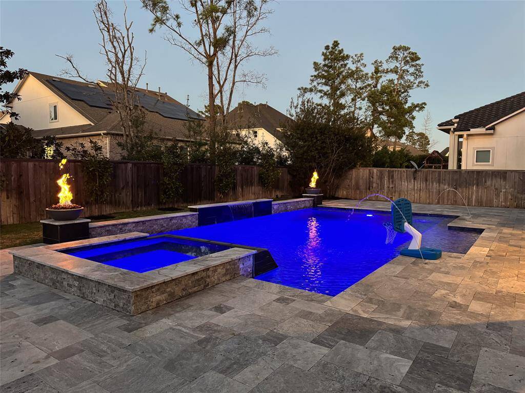 This photo showcases a spacious backyard with a modern swimming pool featuring a built-in spa, decorative fire bowls, and a water slide. The area is surrounded by a wooden fence and landscaped with trees, providing privacy and a serene atmosphere. Perfect for entertaining and relaxation.
