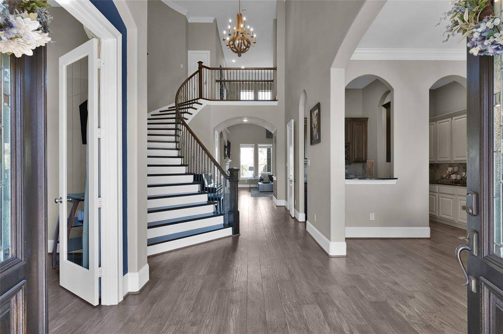 This elegant entryway features a grand staircase with dark wood railing, high ceilings, and a chandelier. The open floor plan leads to a bright living area and a view into the kitchen with arched doorways.