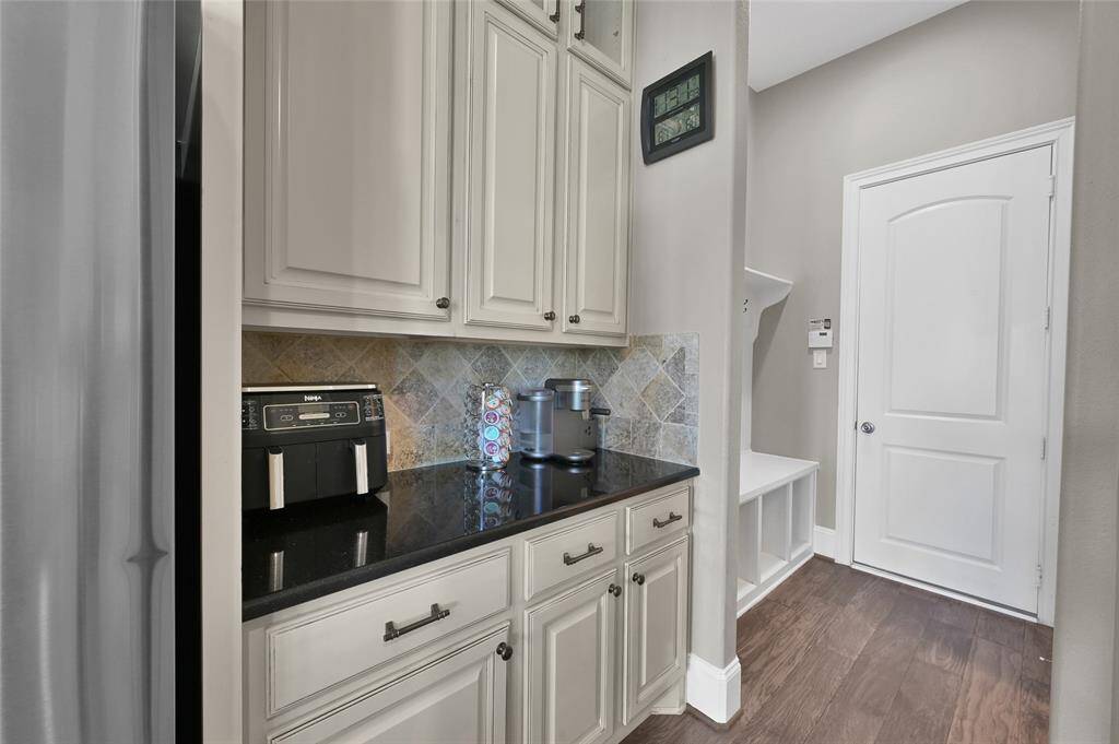 This photo shows a compact kitchen nook with white cabinets, granite countertops and tiled backsplash.  The room has nuetral walls, wood flooring, and a door leading to the garage.  You can see the mudroom area which adds convience for dropping off of backpacks, shoes, etc.