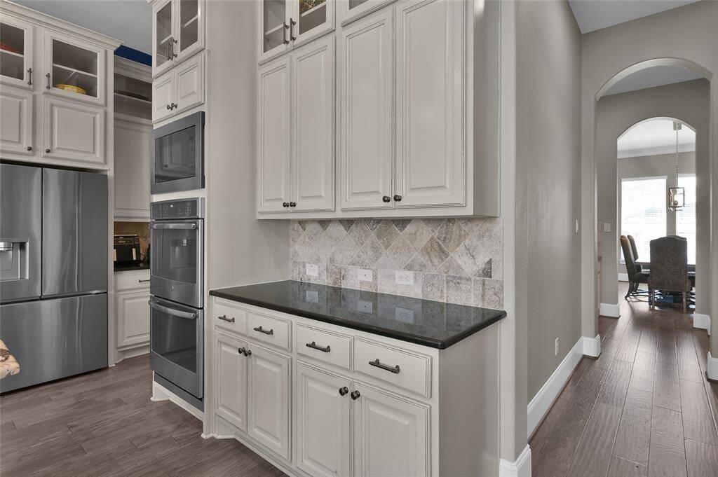 This kitchen features modern double oven stainless steel appliances, elegant cream  cabinetry, a sleek black countertop, and a stylish tile backsplash. The space opens into a bright dining area through an arched doorway, complemented by wood flooring for a warm, inviting feel.