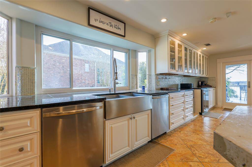 This kitchen features modern high-end stainless steel appliances, including a farmhouse sink and TWO dishwashers (Dacor and Bosch). It has elegant cabinetry with glass fronts, ample counter space, and large windows that provide natural light. The tile flooring and french doors open to expansive deck overlooking the pool and water, enhancing the space's functionality and appeal.