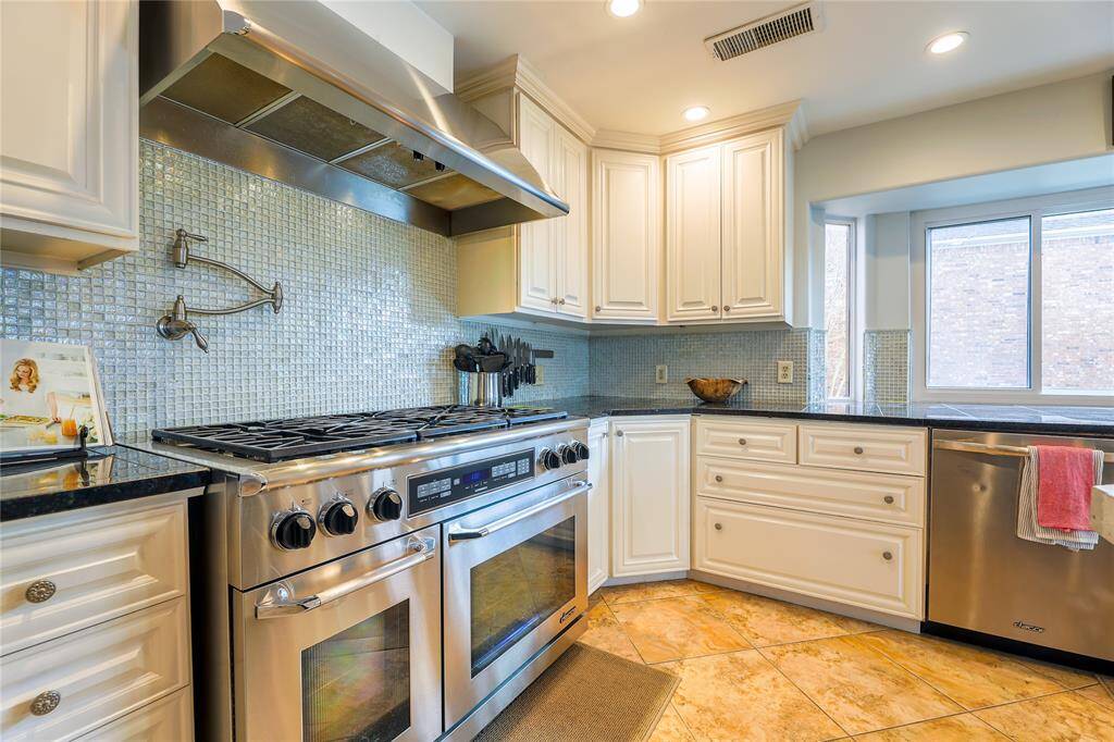 This kitchen features spacious granite countertops, white cabinetry with glass-front doors, and a high-end Dacor gas stove and oven. The room is well-lit with recessed lighting and boasts a modern tile backsplash. A large window fills the space with natural light, enhancing its bright, open feel.