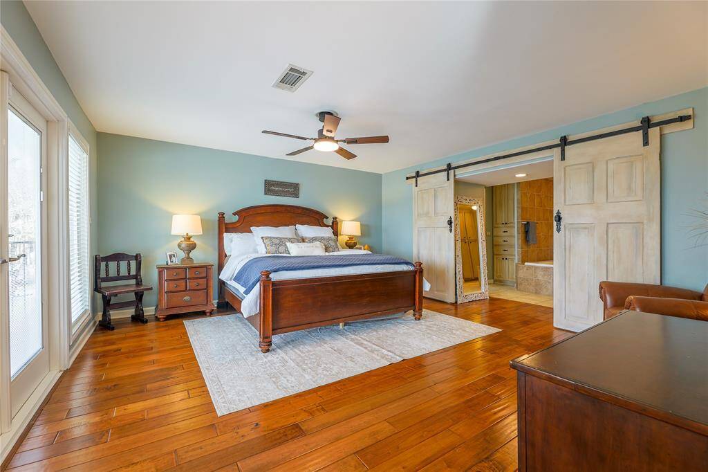 This spacious and cozy primary bedroom features hardwood floors and soft blue walls. It includes a ceiling fan, sliding barn doors leading to a bathroom, and ample natural lighting.