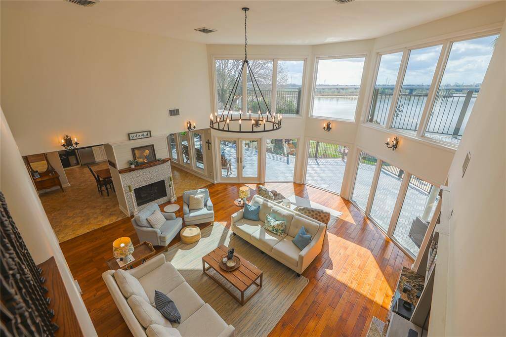 This spacious living room features high ceilings, a wall of large windows offering stunning water views, and abundant natural light. It includes a cozy seating area, a modern fireplace, and elegant wood flooring, creating a warm and inviting atmosphere.
