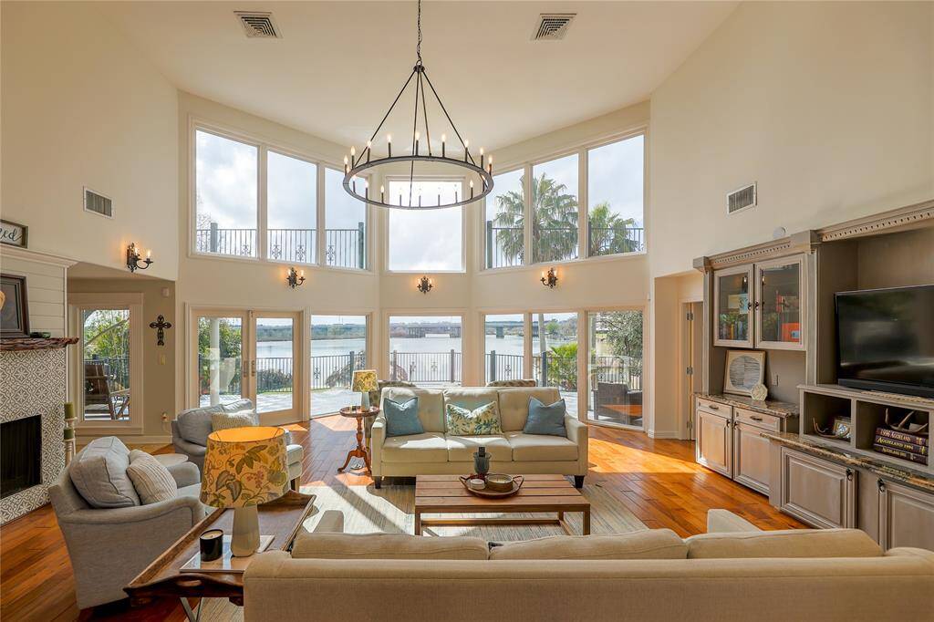 This spacious living room features high ceilings, large windows with water views, and abundant natural light. It includes elegant fixtures, a cozy fireplace, and built-in shelving, creating an inviting and stylish atmosphere perfect for relaxation and entertainment.