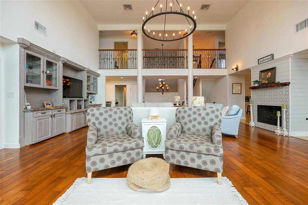 This spacious living room features high ceilings, warm wood flooring, and a cozy fireplace. It includes a large built-in entertainment center and elegant chandelier lighting. The open layout provides a welcoming atmosphere, ideal for entertaining or family gatherings.