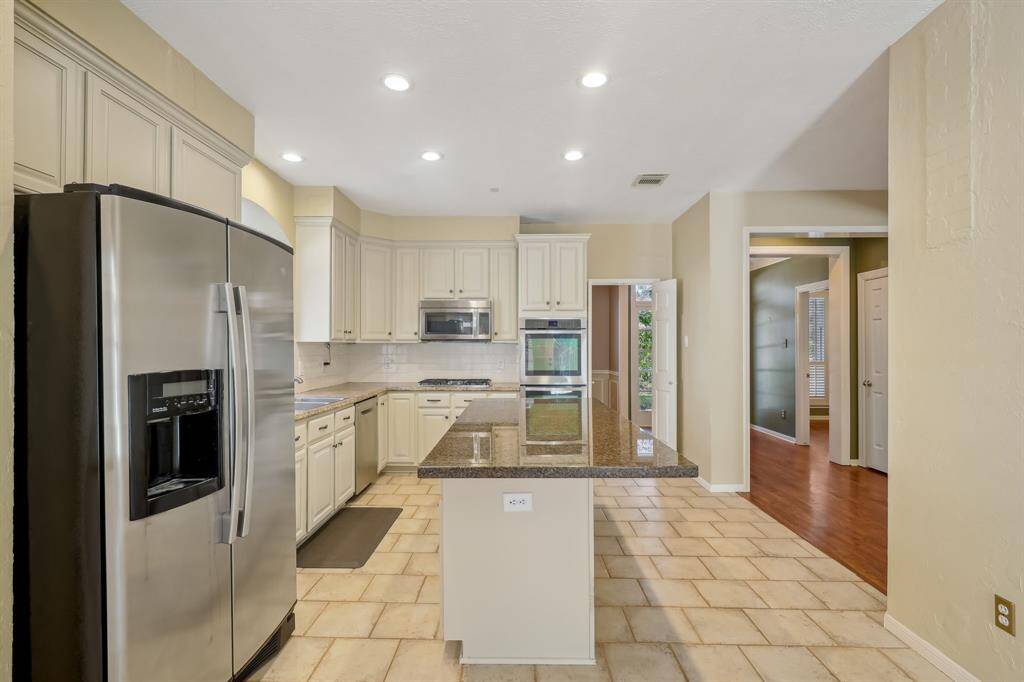 Modern kitchen with stainless steel appliances, granite countertops, and ample cabinetry. Features an island and tile flooring, leading to an open layout with hardwood floors visible in the adjacent room.