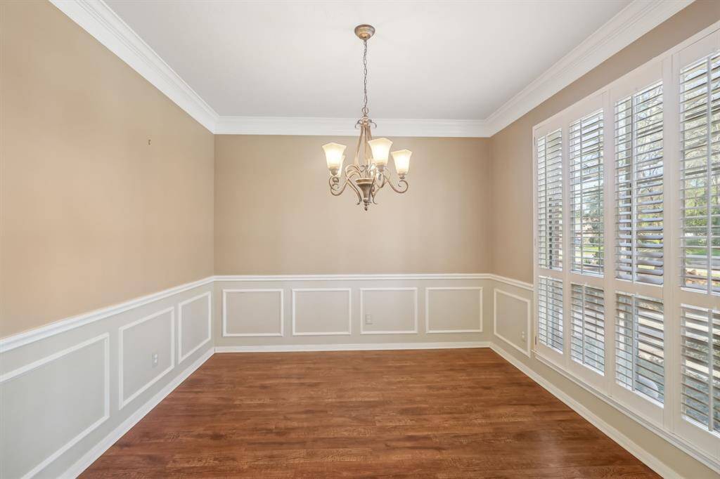This dining room features elegant wainscoting, neutral walls, and a central chandelier. It has large windows with shutters, allowing abundant natural light and showcasing the hardwood floors.