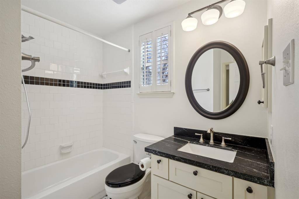 This is a modern bathroom with a white subway-tiled shower and a black accent strip. It features a black countertop with a white sink, a round mirror, and a three-light fixture above. The window has shutters for privacy.