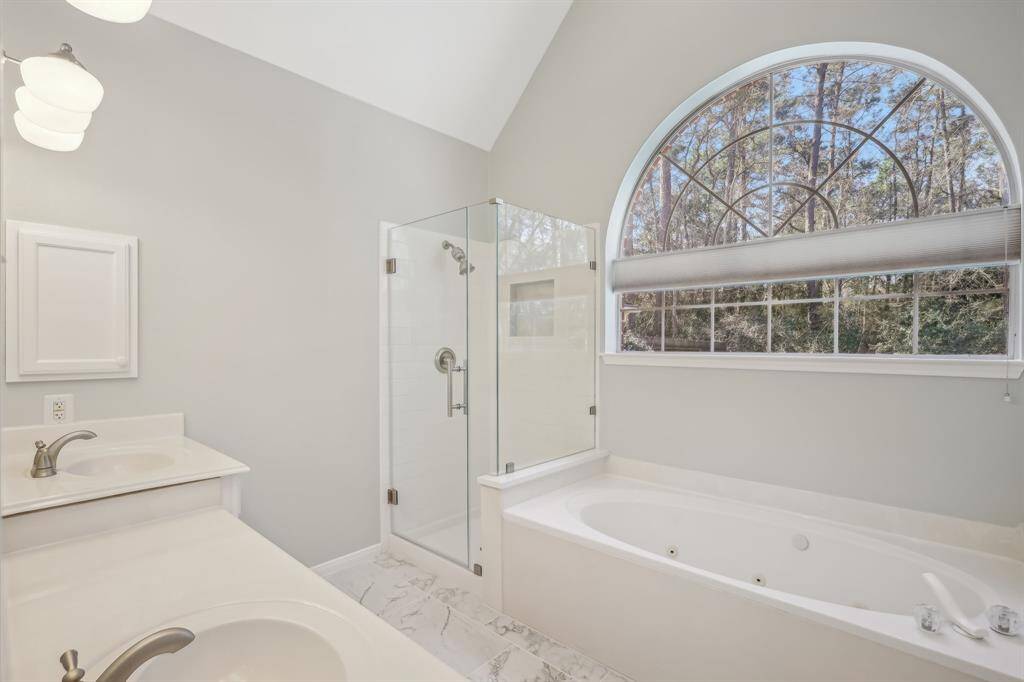 This bright bathroom features a large soaking tub beneath an arched window with a scenic view, a glass-enclosed shower, and dual sinks with modern fixtures, creating a relaxing and elegant space.