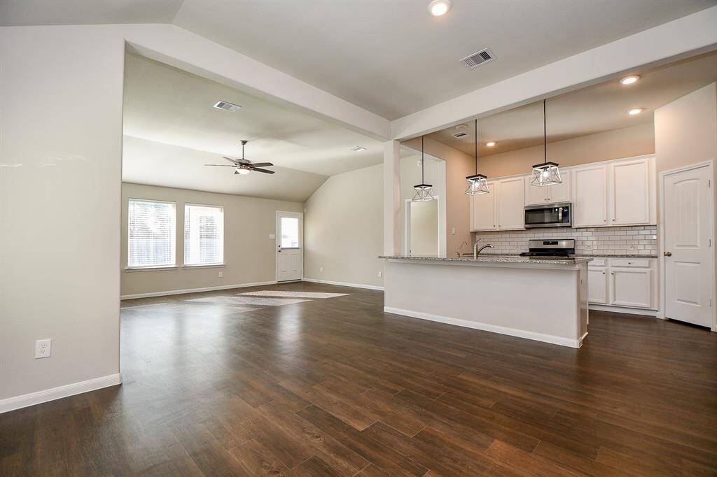 This photo showcases a spacious, open-concept living area with hardwood floors and a modern kitchen. The kitchen features white cabinetry, a large island with granite countertops, and stylish pendant lighting. Large windows provide ample natural light, and there's a ceiling fan for comfort.
