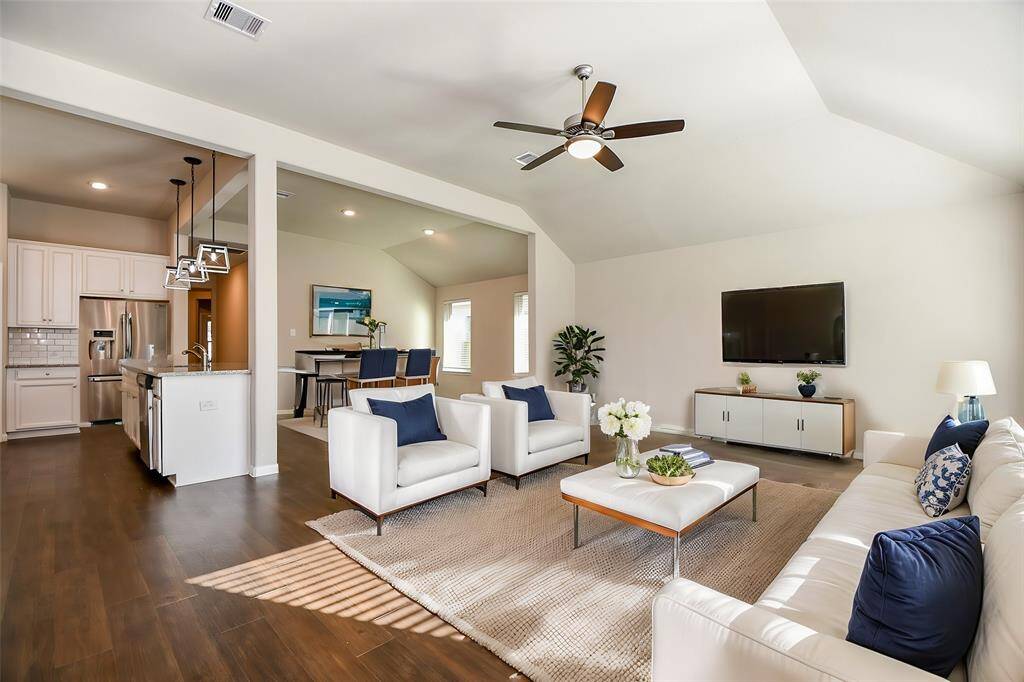 This spacious open-concept living area features a vaulted ceiling with a ceiling fan, hardwood flooring, and abundant natural light. It seamlessly connects to a modern kitchen with white cabinetry, granite countertops, and stylish pendant lighting. Virtually Staged