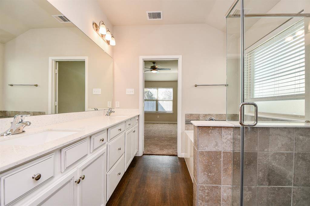 This photo showcases a modern bathroom featuring a double vanity with ample storage, a glass-enclosed shower, and tile details. The space is well-lit with natural light from a large window and has a view into a cozy carpeted bedroom.