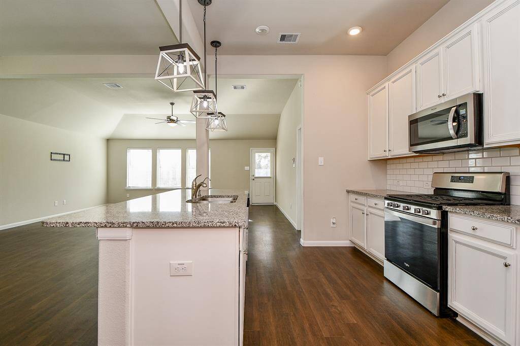 This photo shows an open-concept kitchen and living area with modern appliances, granite countertops, and hardwood floors. The kitchen features white cabinetry, a subway tile backsplash, and pendant lighting over the island. The living space is bright with large windows and a ceiling fan.